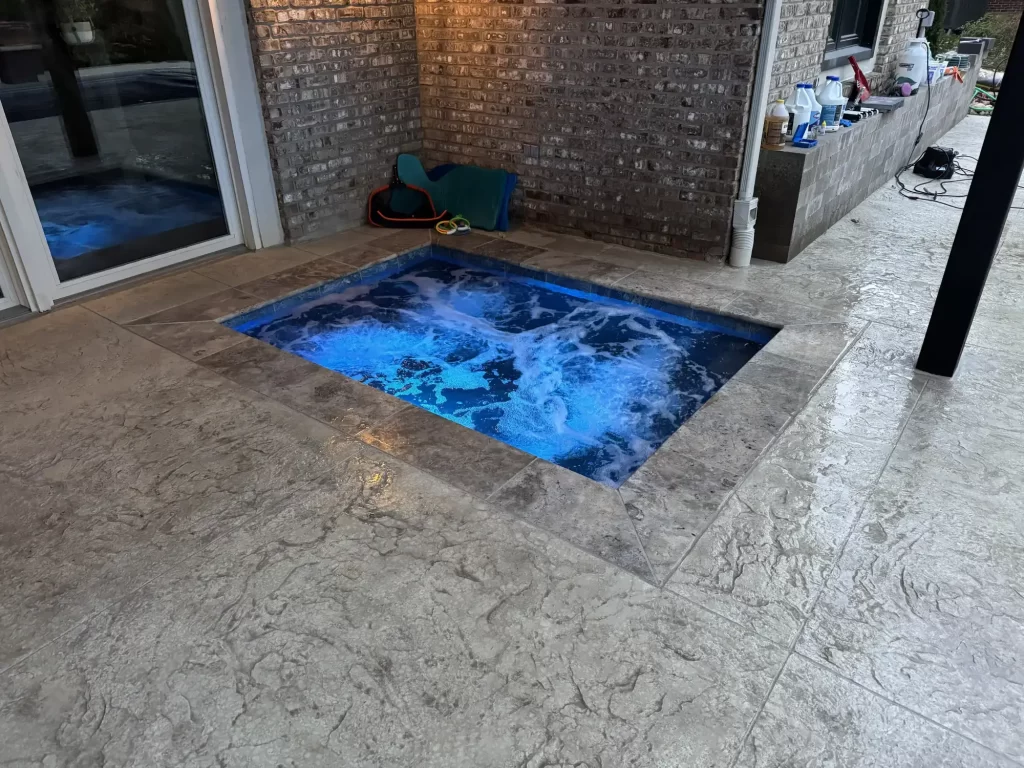 Small square spa set into a stamped concrete patio beside a brick wall