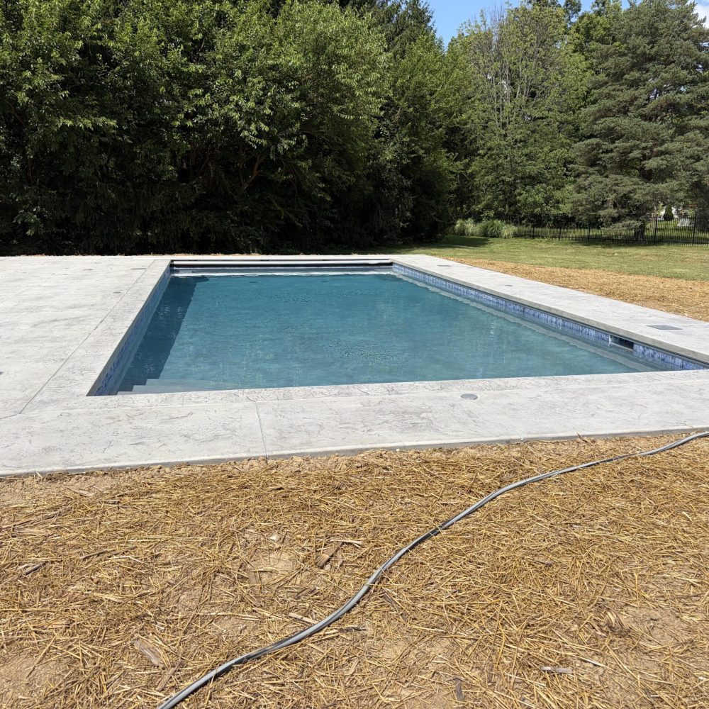 Angled view of a modern rectangular swimming pool with an automatic cover track, textured concrete deck, and brick home backdrop