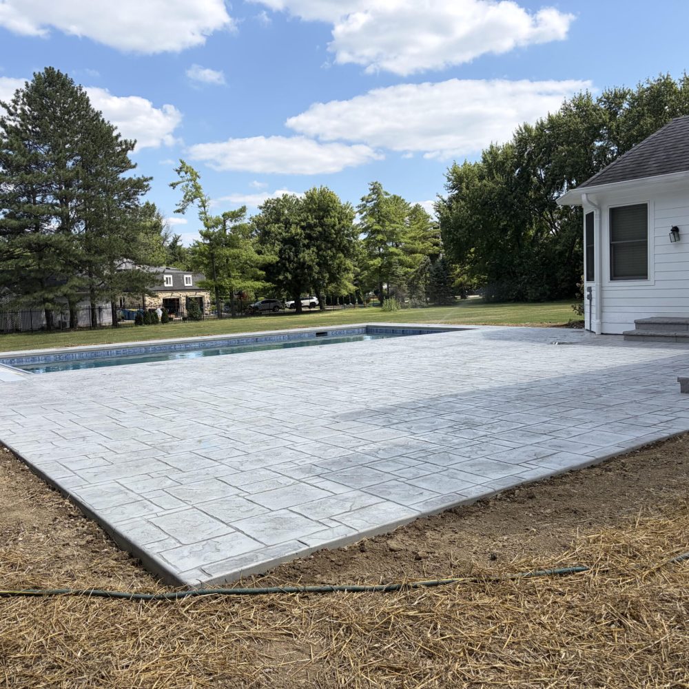 Rectangular backyard pool with built-in entry steps, automatic pool cover, and spacious stamped concrete deck surrounded by mature trees